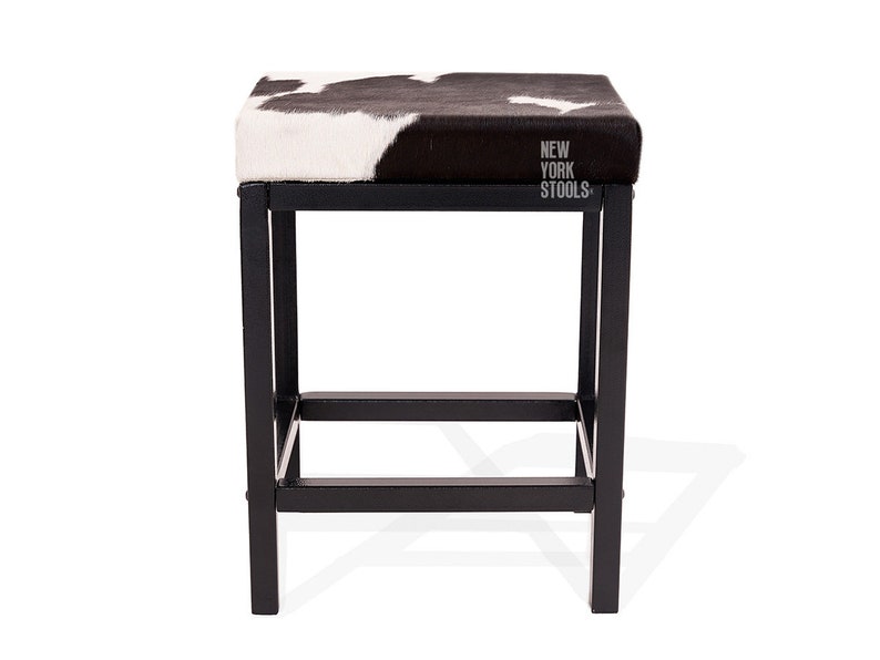 Puede incluir: Un taburete cuadrado con un asiento de patr&oacute;n de piel de vaca en blanco y negro y una estructura de metal negro. El asiento presenta el texto NEW YORK STOOLS impreso en blanco en el lado derecho. La estructura tiene cuatro patas rectas.