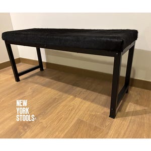 Puede incluir: Un banco rectangular con un asiento negro texturizado y una estructura de metal negro con cuatro patas inclinadas. El texto NEW YORK STOOLS está impreso en blanco en la parte inferior izquierda del marco. El banco tiene un diseño estructural limpio.