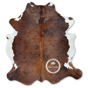 以下が含まれることがあります： 茶色と白の牛革の敷物で、自然な斑点模様があります。敷物は約1.5メートル×2.1メートルです。