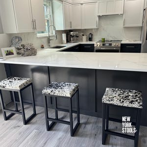 Può includere: Tre sgabelli da bar in metallo nero con sedili a fantasia pelle di mucca sono posizionati davanti a un'isola cucina. La cucina presenta mobili bianchi, un piano di lavoro in marmo e un frigorifero in acciaio inossidabile. Il testo "NEW YORK STOOLS" è visibile.