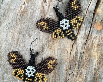 May include: A pair of beaded earrings shaped like moths with brown, gold, black and white beads. The moths have a skull and crossbones design on their bodies.