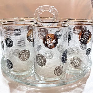 May include: A set of clear glass tumblers with gold rims and a decorative pattern of various international coins, arranged on a matching circular glass serving tray with a central handle. The coins feature detailed illustrations and text.