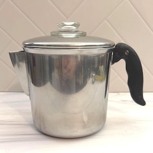 May include: A polished stainless steel percolator coffee pot featuring a cylindrical body, a side-mounted black ergonomic handle, and a matching stainless steel lid topped with a clear glass knob. The spout is integrated into the side.