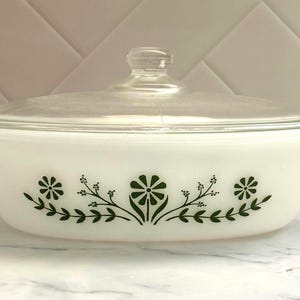 Puede incluir: Una fuente de horno ovalada de vidrio opalino blanco con tapa de vidrio transparente. El exterior presenta un patrón floral verde compuesto por margaritas estilizadas y ramas con hojas centradas en el lateral. Tiene asas integradas.