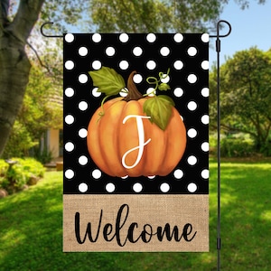May include: A rectangular garden flag featuring a black background with white polka dots. A large orange pumpkin with green leaves and a vine is centered, displaying a white letter J. A tan burlap-textured band at the bottom reads Welcome in black.