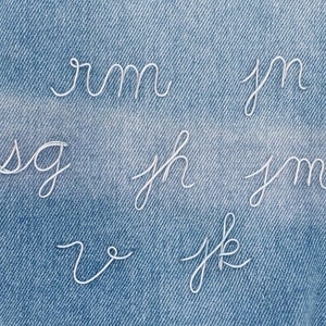 Peut inclure: Lettres cursives en fil blanc sur un fond en denim bleu. Les lettres incluent 'rm', 'jn', 'sg', 'jh', 'jm', 'v' et 'jk'. Les lettres sont de style manuscrit, et le denim a un aspect texturé.