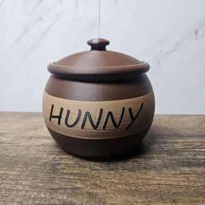 May include: A brown and tan ceramic honey pot with a lid. The word "HUNNY" is written in black letters across the center of the pot. The pot is sitting on a wooden surface.