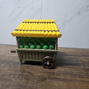 May include: A miniature wooden cart with a yellow roof and a brown wheel. The cart is filled with green produce, possibly cabbages or similar vegetables. The cart has a handle and is designed to resemble a market stall.