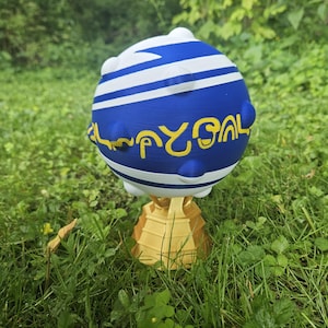 May include: A 3D-printed decorative sphere with a blue and white striped design and yellow lettering. The sphere sits atop a gold-colored, pyramid-shaped base. The object is placed in a grassy outdoor setting.