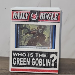May include: A white card holder with the text "DAILY BUGLE" and "WHO IS THE GREEN GOBLIN?" in black and red. The holder contains a card with a fantasy illustration. The holder is on a wooden surface.