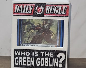 May include: A white card holder with the text "DAILY BUGLE" and "WHO IS THE GREEN GOBLIN?" in black and red. The holder contains a card with a fantasy illustration. The holder is on a wooden surface.