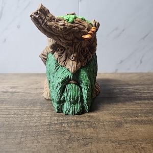 May include: A whimsical, handcrafted figurine of a forest spirit. The sculpture features a detailed, textured brown bark-like head with green moss and orange mushrooms. It has a green, textured beard, and is set on a wooden surface.
