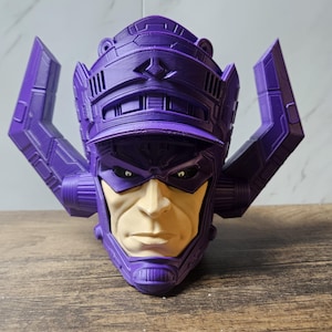 May include: A 3D printed bust of the character Galactus, featuring a prominent purple helmet with large, angular side protrusions. The face is painted in a light beige tone with dark, intense eyes, and the helmet displays detailed mechanical textures.