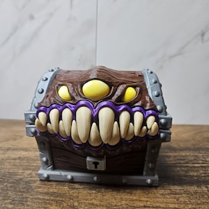May include: A brown treasure chest with a monster face design. The chest features three yellow eyes, a purple-lined mouth filled with white teeth, and gray metal accents. The chest has a keyhole and is set on a wooden surface.