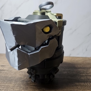 May include: A detailed, dark gray and black prop resembling a stylized grenade. The prop features a textured, angular design with a yellow eye-like detail and a ring on top. The object is set against a neutral background.