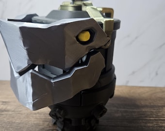 May include: A detailed, dark gray and black prop resembling a stylized grenade. The prop features a textured, angular design with a yellow eye-like detail and a ring on top. The object is set against a neutral background.