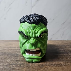 May include: A 3D-printed planter shaped like the head of the Hulk, featuring bright green skin, a furrowed brow, black hair, and an open mouth showing white teeth. The surface displays horizontal layer lines characteristic of additive manufacturing.