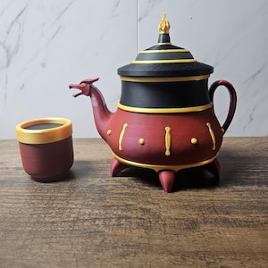 May include: A decorative teapot and cup set. The teapot is red with black and gold accents, featuring a dragon-like spout and a flame-shaped finial. The cup is a matching red with a gold rim. The set is displayed on a wooden surface.