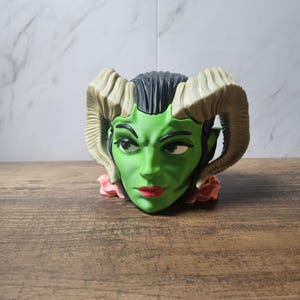May include: A green-skinned, horned face planter with red lips and black hair. The planter has large, curved horns and a pink floral base. The face has a stern expression and is set against a wooden surface.