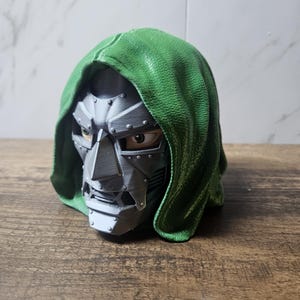 May include: A three-dimensional bust of a character wearing a metallic silver mask with visible rivets and a textured green hood. The mask features a sharp nose piece, a structured jawline, and visible eyes peering through the eye openings.