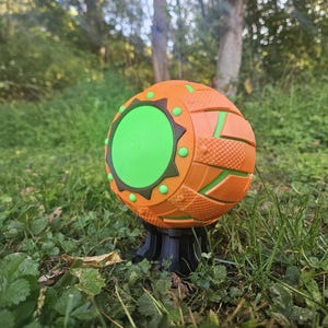 May include: An orange and green spherical toy with a textured surface and a circular green center. The toy has a black base and is set against a backdrop of green grass and trees.