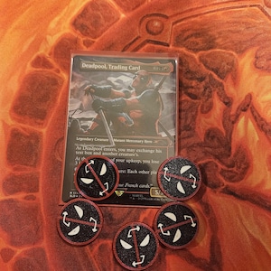 May include: A Deadpool trading card with a black and red character illustration. The card is surrounded by six black and red tokens with a white skull and crossbones design.