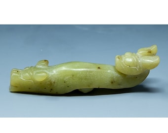 Puede incluir: Una escultura de dragón de jade verde con detalles intrincados. El dragón está tallado en posición reclinada con la cabeza girada hacia un lado. La escultura está hecha de jade liso y pulido.