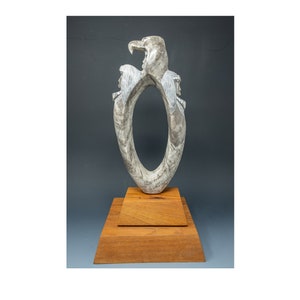 May include: A white stone sculpture of three figures, two with their heads turned to the side and one with its head facing forward, forming a circle. The sculpture is on a wooden base.