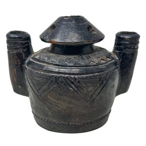 Puede incluir: Un recipiente de cerámica marrón oscuro de aspecto antiguo, con tapa y dos picos. El recipiente tiene patrones geométricos grabados y un aspecto desgastado, lo que sugiere edad y uso. La tapa tiene varios agujeros pequeños.
