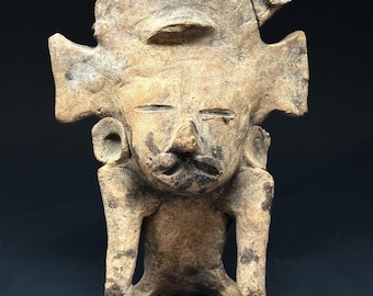 May include: A tan-colored, ancient-looking figurine with a stylized human form. The figure has a large headdress, prominent ears, and a detailed face with a mustache. The arms are bent, and the figure appears to be seated. The sculpture has dark markings.