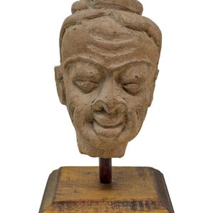 Könnte beinhalten: Eine hellbraune, verwitterte Terrakotta-Skulptur eines menschlichen Kopfes mit gelassenem Ausdruck, geschlossenen Augen und einem strukturierten Haarknoten. Der Kopf ist auf einem dunkelbraunen Stab auf einem quadratischen Holzsockel montiert.