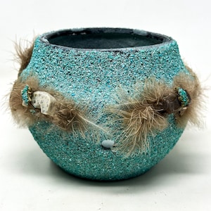 Puede incluir: Una maceta de cerámica turquesa texturizada con un borde oscuro. Está adornada con plumas marrones y pequeños elementos decorativos. La maceta tiene forma redondeada y una superficie rugosa y granulada.