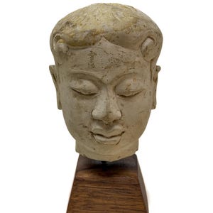 Puede incluir: Una escultura de piedra de color blanco roto, desgastada, que representa una cabeza humana con los ojos cerrados y el cabello estilizado, montada sobre una base de madera trapezoidal de color marrón oscuro. La piedra muestra signos de edad.