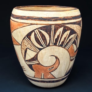 May include: A beige ceramic vase with a rounded shape and a wide opening. The vase features intricate brown and orange geometric designs, including a spiral and stylized shapes. The design is symmetrical and covers the majority of the vase's surface.