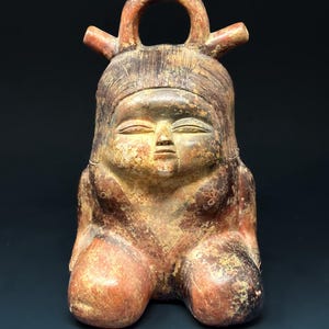 May include: A weathered, terracotta-colored ceramic figure of a seated person with a serene expression. The figure has a rounded body, with legs bent and knees drawn up. The head features a loop and two small protrusions, and the face has closed eyes and a small mouth.