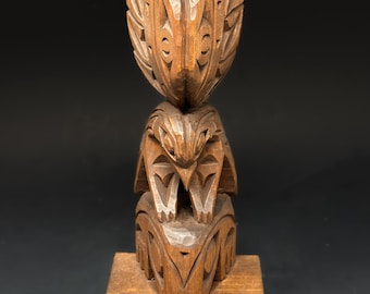 May include: A wooden totem pole with three figures carved in a traditional style. The top figure is a bird with outstretched wings, the middle figure is a bear, and the bottom figure is a stylized human face. The totem pole is mounted on a wooden base.