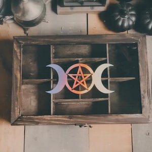 May include: A dark wooden display case with multiple compartments. The case features a central design of a pentagram within a circle, flanked by crescent moons. The design has a gradient of colors, including orange, pink, and blue. The case is likely for displaying small items.