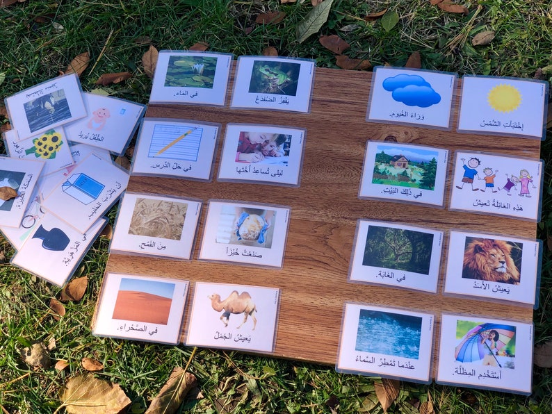 May include: A set of colorful flashcards with images and Arabic text. The cards feature various topics, including animals, nature, and daily activities. The cards are arranged on a wooden surface.