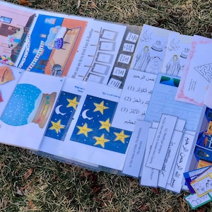 Puede incluir: Una colección de materiales educativos coloridos, que incluyen páginas ilustradas, pegatinas en forma de corazón y marcadores. Las páginas presentan dibujos de edificios, estrellas y texto en árabe. Los artículos están dispuestos sobre una lámina de plástico transparente.