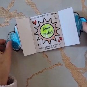 May include: A white card with a sun design and the words "Imam Mahdi" in the center. The card has a cloud shape on each side and text that reads "I shine to show you the way" and "Take a step toward me, start today!"