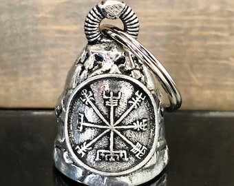 May include: A silver-colored bell-shaped keychain with a detailed, weathered texture. The bell features a circular emblem with a symmetrical design. The top has a ring for attachment, and the background is a blurred wooden surface.