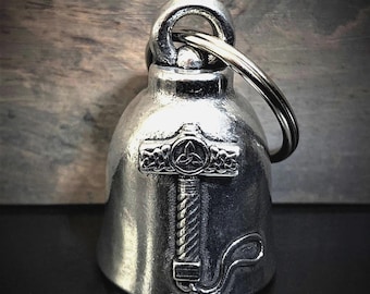 May include: A silver-colored bell with a loop for hanging and a key ring. The bell features an embossed design of a hammer with a Celtic knot symbol. The bell is likely made of metal and has a shiny finish.