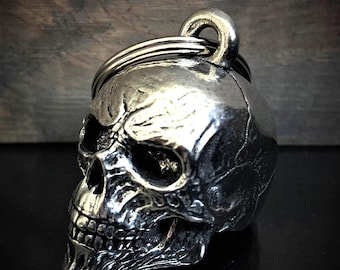 May include: A silver-colored skull-shaped keychain with a ring for attaching keys. The skull has detailed features, including eye sockets, a nose cavity, and teeth. The keychain is made of a shiny metal and reflects light.