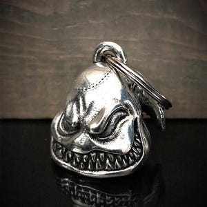 Puede incluir: Una campana plateada con forma de rostro sonriente, con dientes afilados y un anillo para colgar. La campana tiene una superficie texturizada y detallada, probablemente de metal. El diseño evoca un tema de fantasía u horror.