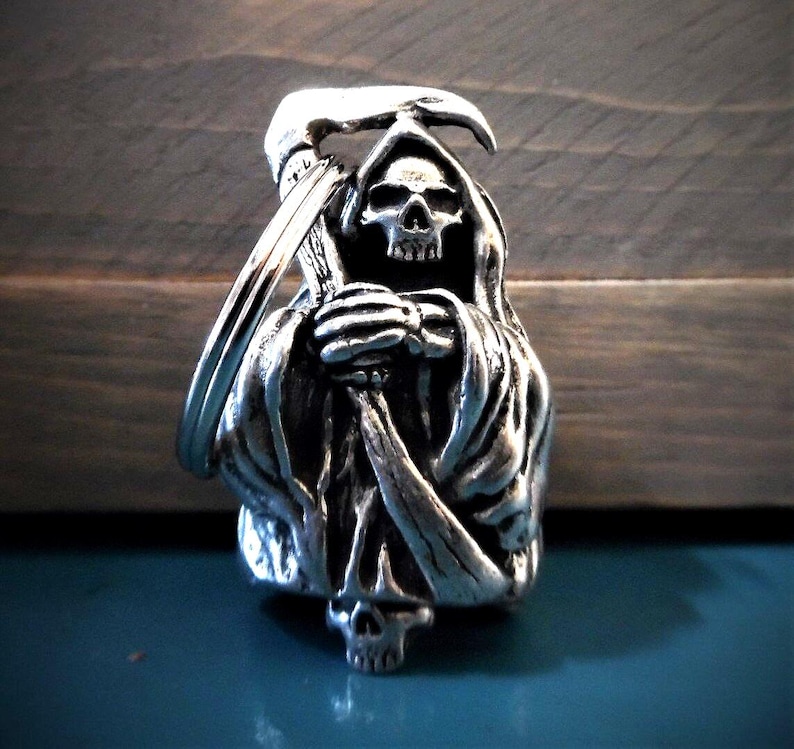 Puede incluir: Llavero plateado con una escultura detallada de la Muerte sosteniendo una guada&ntilde;a. La Muerte lleva una t&uacute;nica con capucha y tiene una cara esquel&eacute;tica.