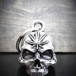 May include: A silver-colored skull-shaped bell with a cannabis leaf design on top. The bell has a ring for hanging and is made of a shiny metal. The skull has detailed features, including eye sockets, a nose cavity, and teeth.