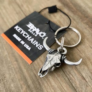 Może przedstawiać: Srebrny brelok do kluczy z czaszką byka z zakrzywionymi rogami i k&oacute;łkiem na klucze. Brelok jest przymocowany do czarnej etykiety z napisem "BRAVO BELLS KEYCHAINS MADE IN USA". Tło to drewniana powierzchnia.