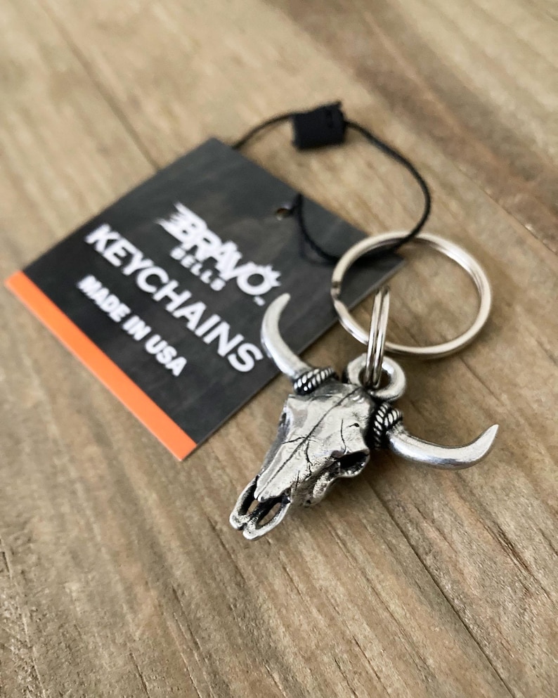 Może przedstawiać: Srebrny brelok do kluczy z czaszką byka z zakrzywionymi rogami i k&oacute;łkiem na klucze. Brelok jest przymocowany do czarnej etykiety z napisem "BRAVO BELLS KEYCHAINS MADE IN USA". Tło to drewniana powierzchnia.