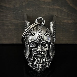 May include: A silver-colored metal keychain featuring a detailed Viking warrior helmet design. The helmet has intricate carvings, a long beard, and a ring for attaching keys. The background is a dark wooden surface.