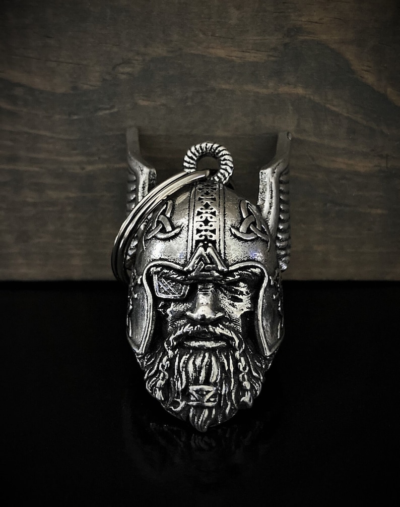 May include: A silver-colored metal keychain featuring a detailed Viking warrior helmet design. The helmet has intricate carvings, a long beard, and a ring for attaching keys. The background is a dark wooden surface.
